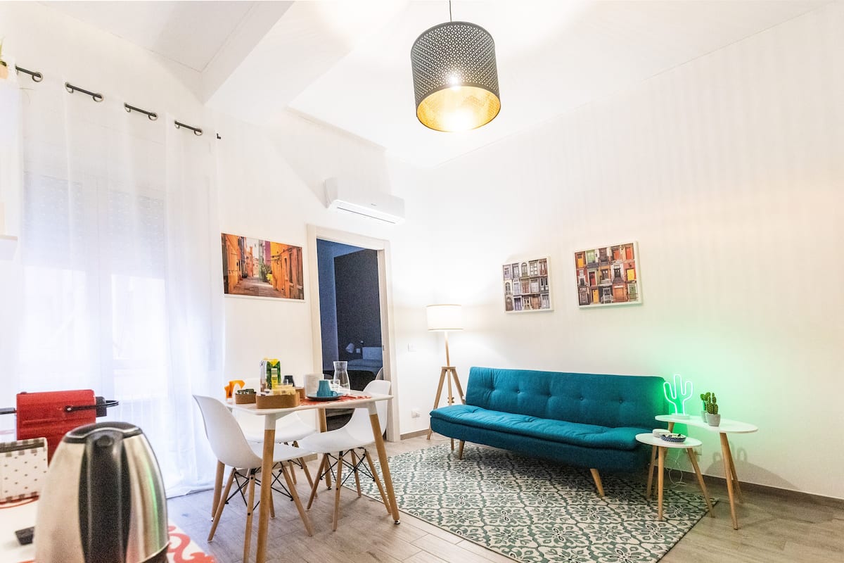 A modern living room with a teal sofa, a dining table with chairs, a coffee machine, and a small side table with a lamp.