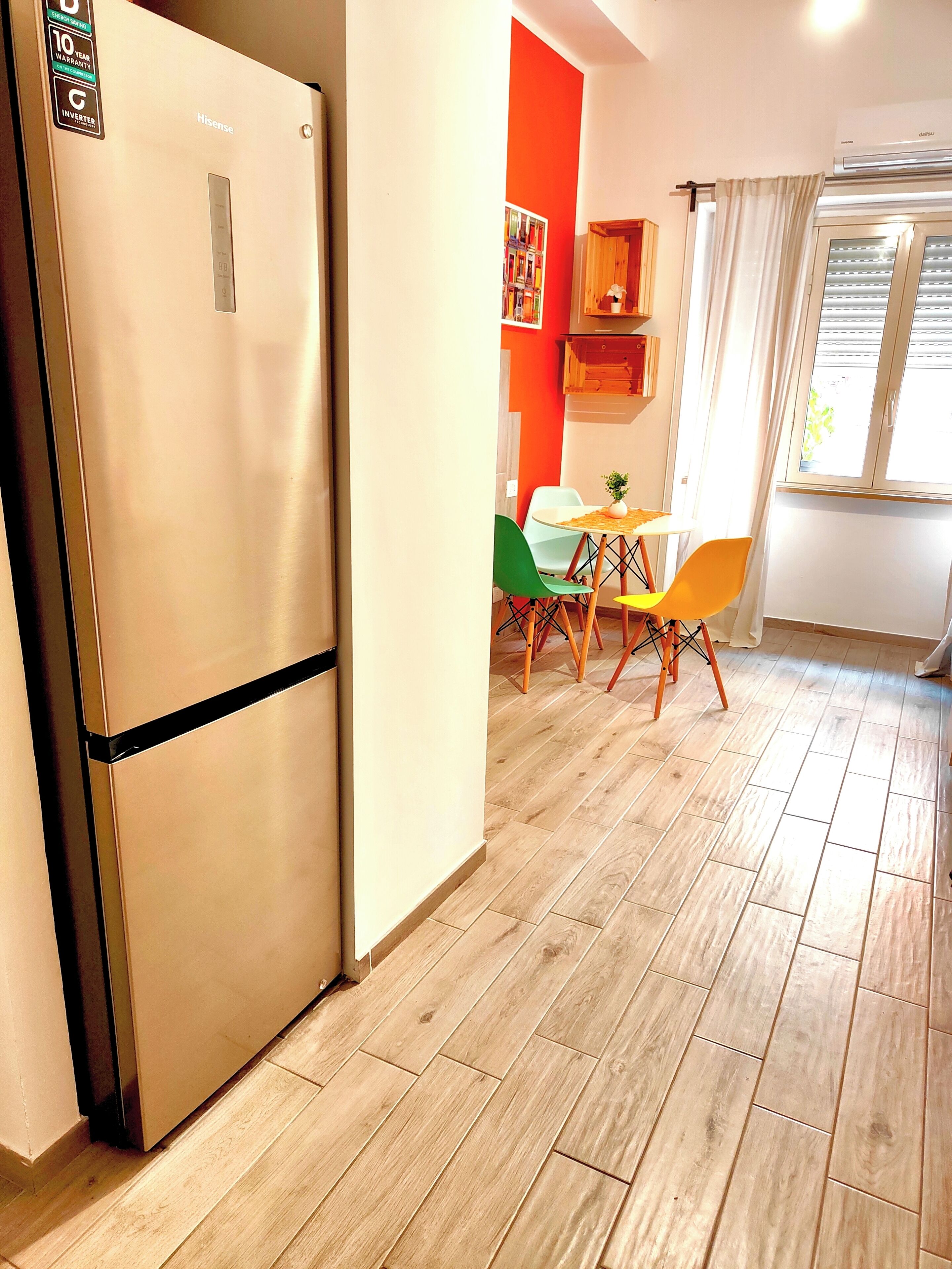 A modern kitchen with a stainless steel refrigerator, wooden flooring, and a dining area with colorful chairs.