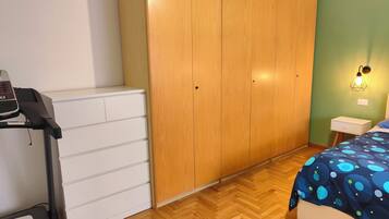 Un dormitorio con un armario de madera, una cinta de correr, una cama con edredón estampado y una mesita de noche con lámpara.