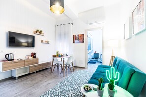 Una sala moderna con un televisor de pantalla plana, una zona de comedor con mesa y sillas, un sofá azul y una decoración de cactus.