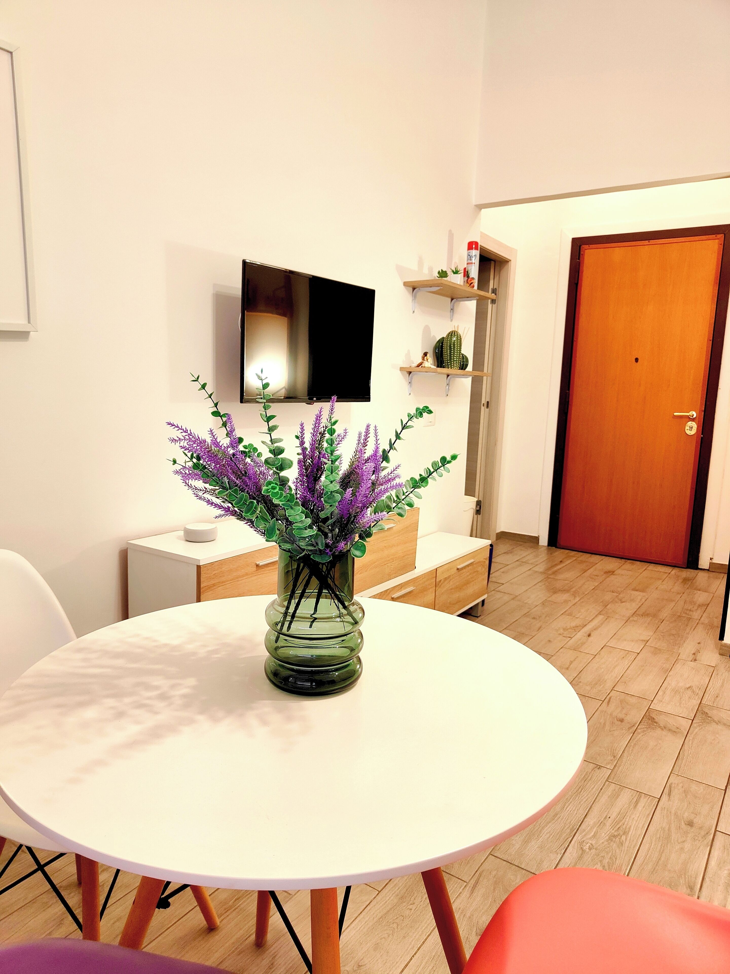 Een moderne woonkamer met een ronde witte tafel, een glazen vaas met paarse bloemen, een flatscreen televisie aan de muur en een houten deur.