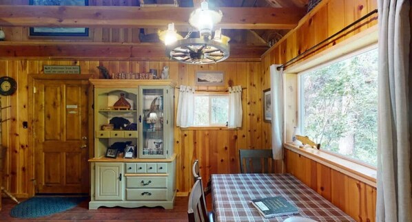 Interior de una cabaña con una mesa de comedor, un armario con puertas de cristal, una ventana con cortinas y una lámpara en el techo.