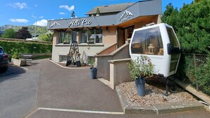 Un hotel llamado "Alti Pic" con un diseño moderno, una escalera que conduce a la entrada y un área de estacionamiento con un automóvil y una camioneta blanca.