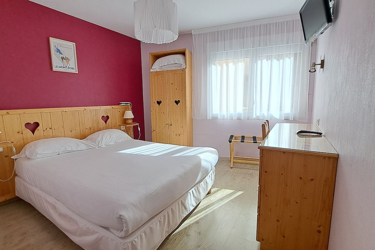 A bedroom with a wooden headboard, a bed with white bedding, a wooden wardrobe, a desk with a chair, and a TV mounted on the wall.