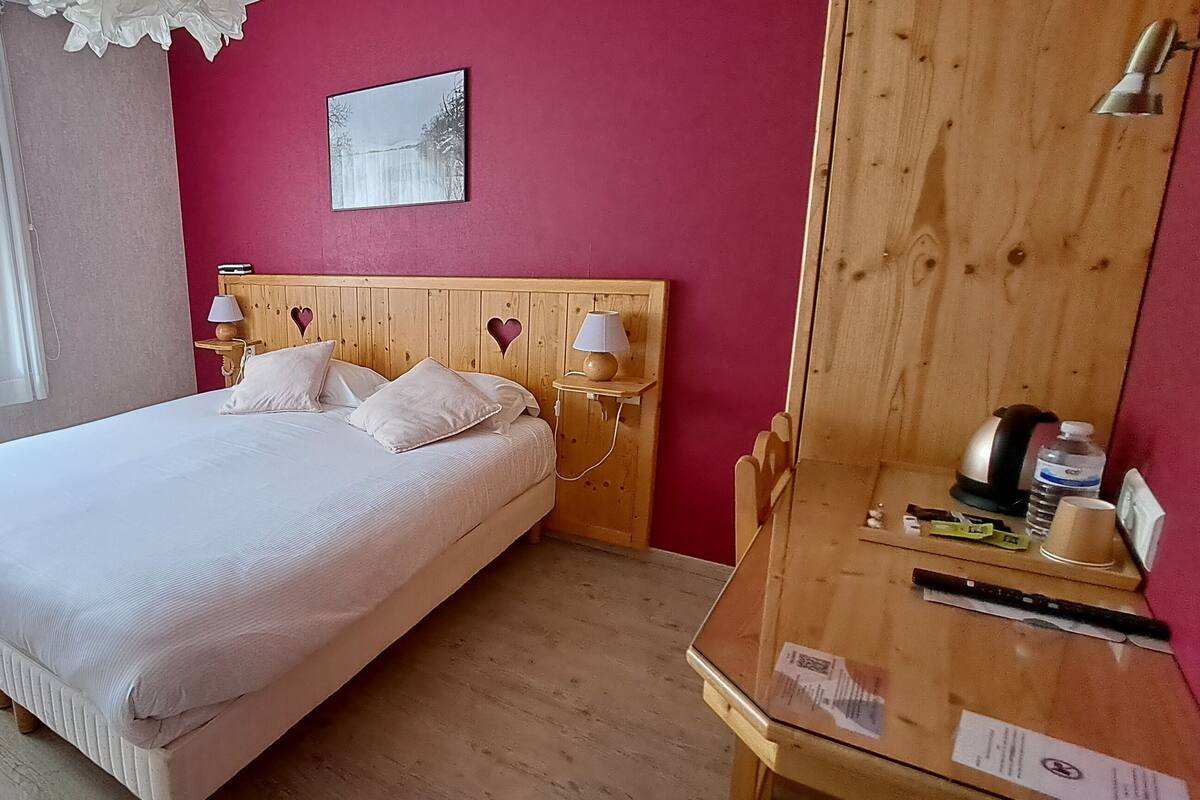 A bedroom with a bed, two bedside tables with lamps, a wooden wardrobe, and a window with sheer curtains.