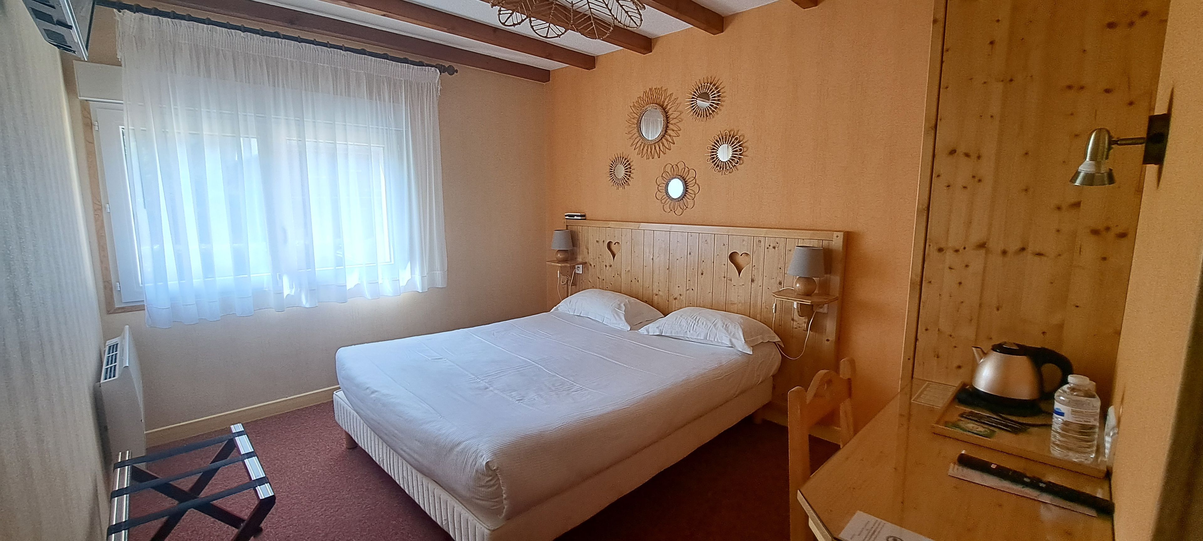 A bedroom with a bed, two bedside lamps, a wooden headboard, a window with sheer curtains, and a small table with a kettle and water bottle.
