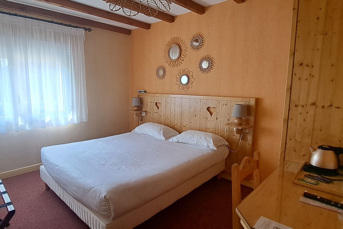 A bedroom with a bed, two bedside lamps, a wooden headboard, a window with sheer curtains, and a small table with a kettle and water bottle.