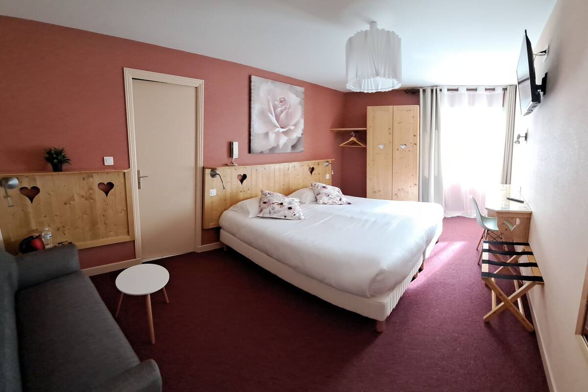 A hotel room with a large bed, a desk, a chair, a sofa, a wardrobe, and a mirror.
