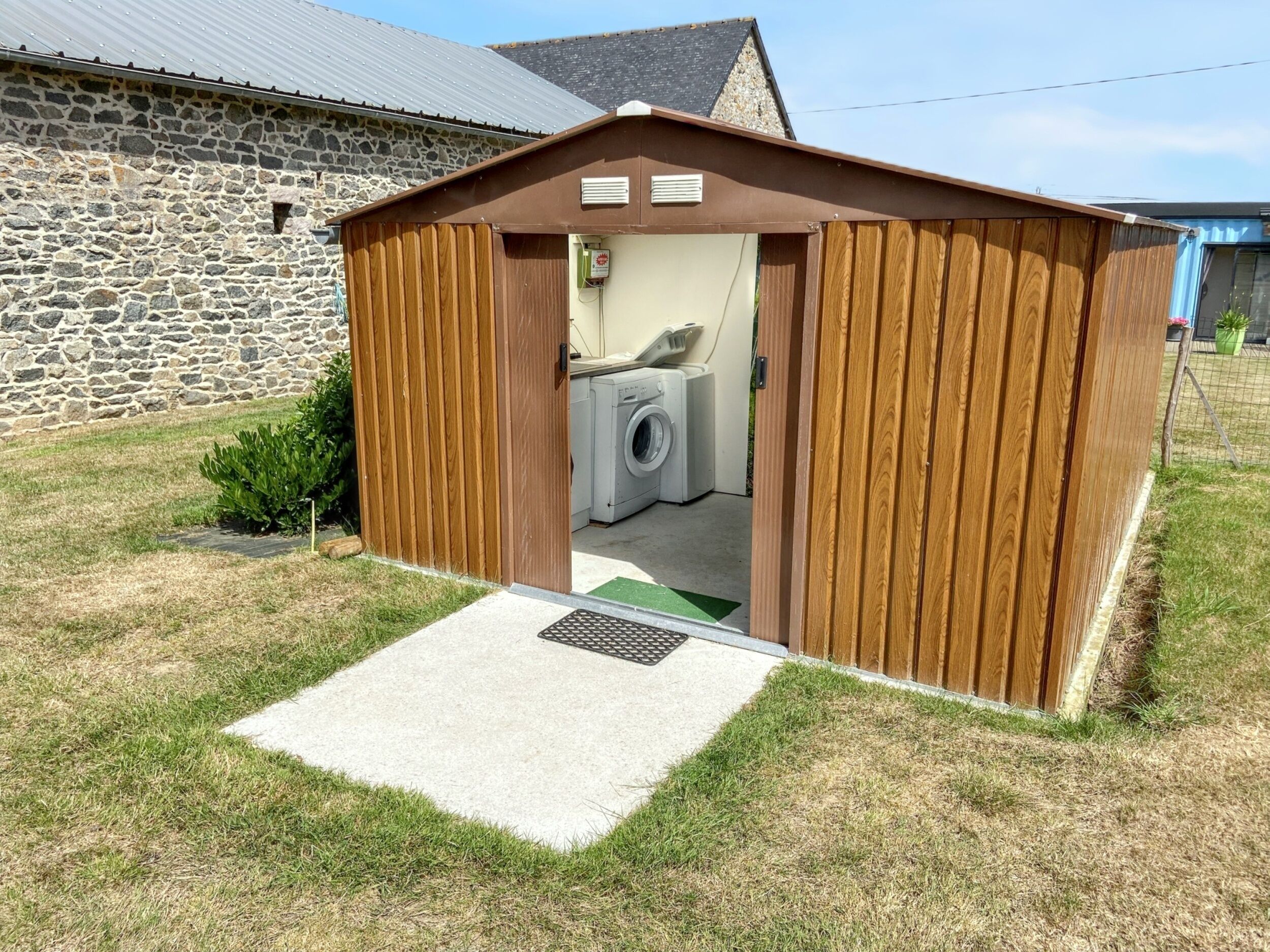 Un petit abri en bois abritant un lave-linge, un mur de pierres en arrière-plan et une cour couverte d’herbe.
