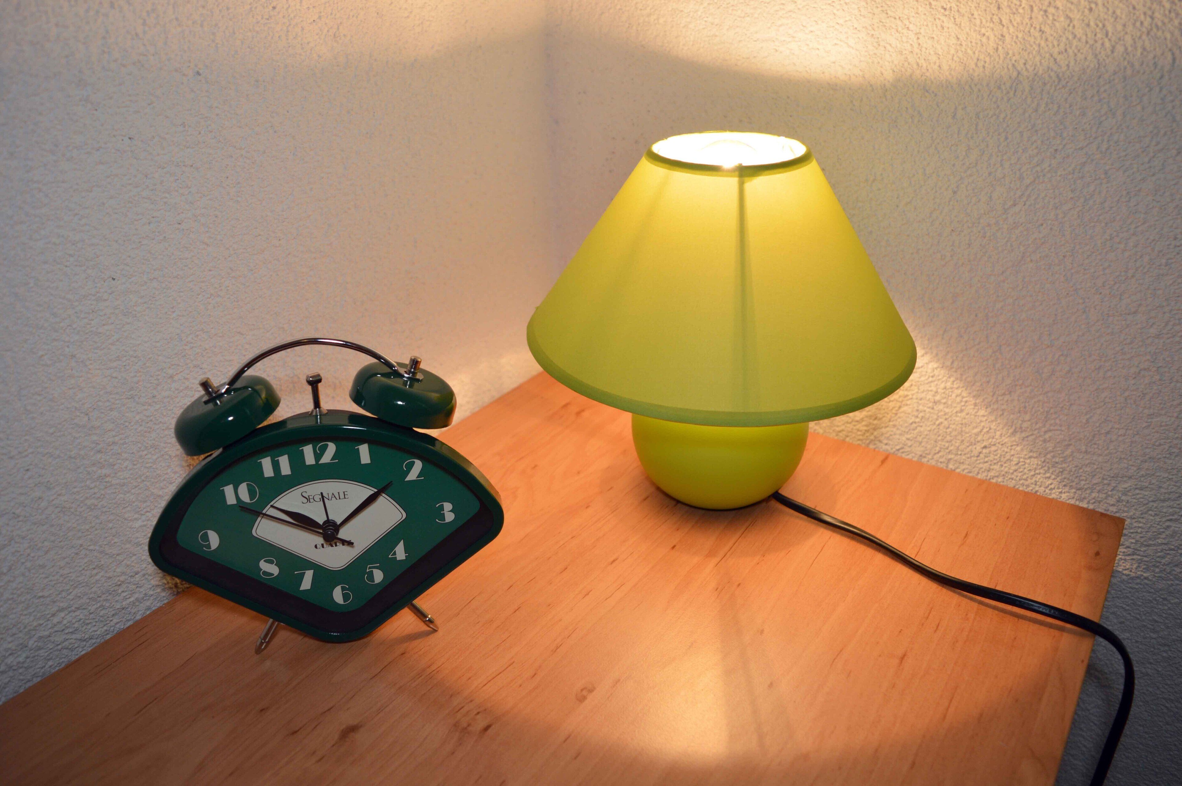 A green analog alarm clock and a yellow table lamp on a wooden table against a white wall.