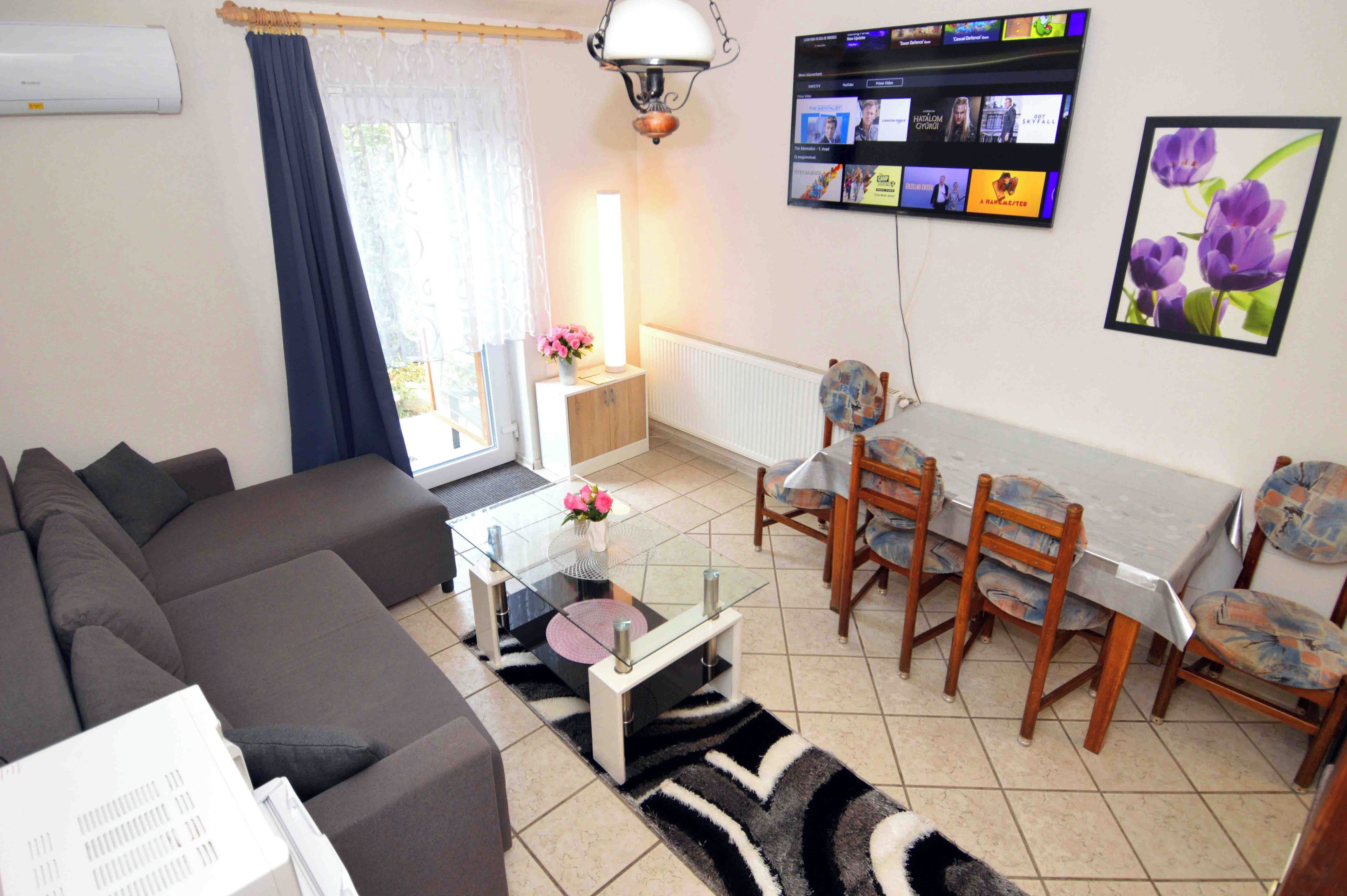 A living room with a sofa, a dining table with chairs, a TV mounted on the wall, and a window with curtains.