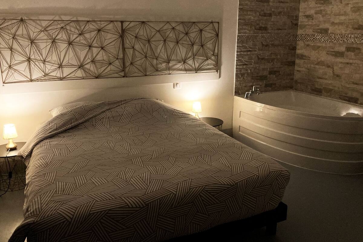 Une chambre à coucher comprenant un lit, un luminaire géométrique fixé au mur, une table de chevet avec une lampe et une baignoire.