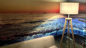 A room with a sofa and a floor lamp, a wall mural depicting a seascape, and a patterned throw blanket. - Desir'Add (La Clisse)