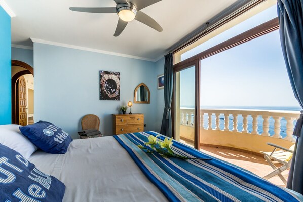 Un dormitorio con cama, cómoda, silla, ventilador de techo, ventana grande con vista al mar y balcón con barandilla.