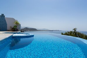 Una piscina infinita con agua azul cristalina, un muro de piedra y vistas al mar y montañas lejanas.
