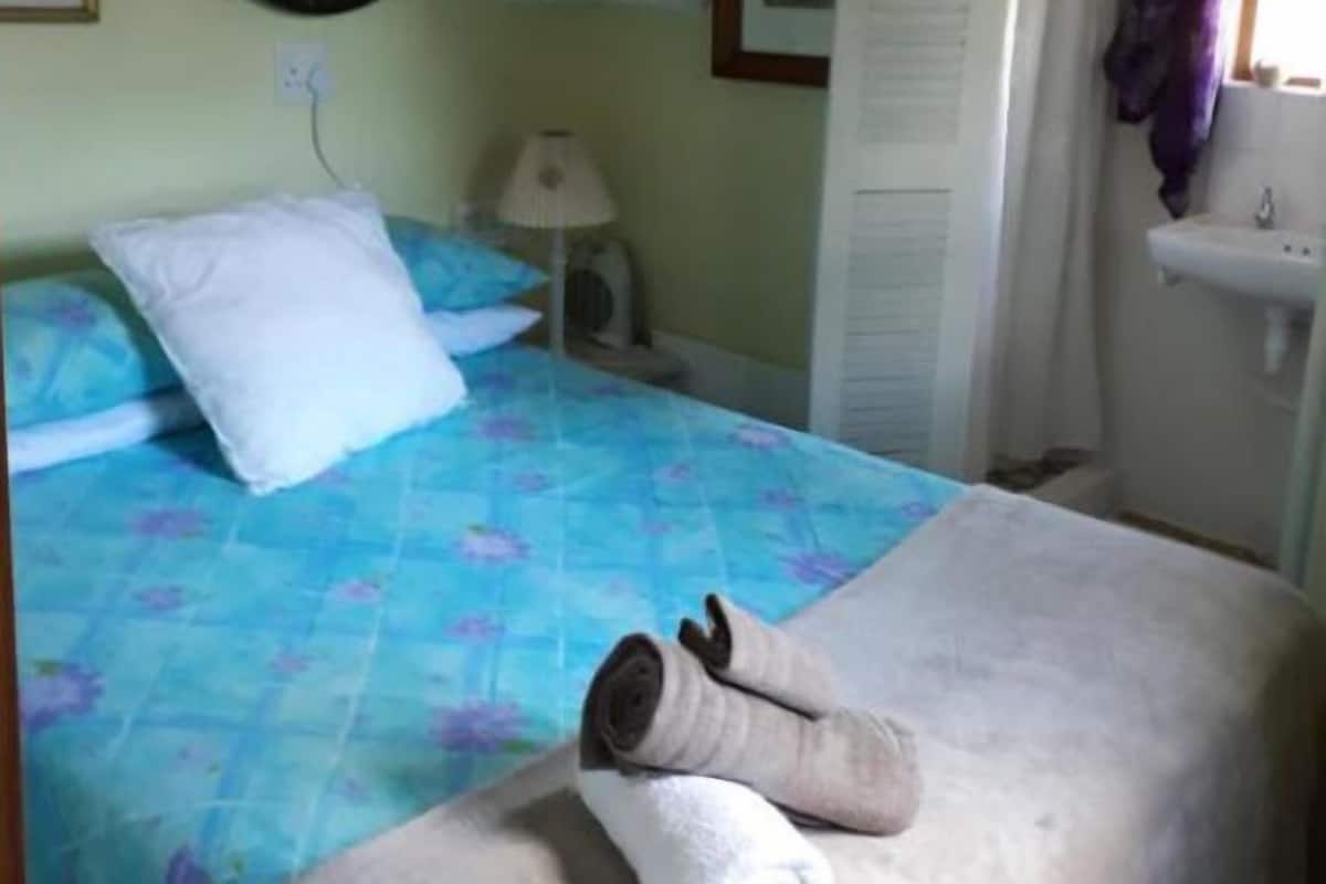 A bedroom with a bed, a bedside lamp, a shelf with decorative items, a window with curtains, and a bathroom with a sink.