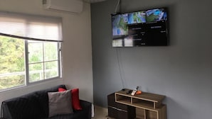 A modern living room with a flat-screen TV mounted on the wall, a dark-colored sofa with red cushions, a wooden side table, and a window with blinds.