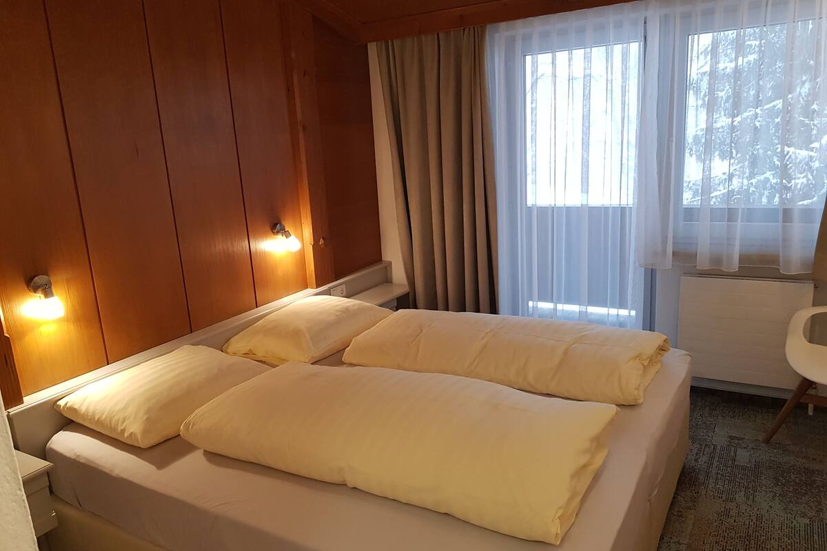 A hotel room with a bed, wooden walls, a window with curtains, and a radiator.