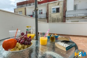 A table with a glass of orange juice, a glass of water, a bowl of grapes, a book, and a bottle of olive oil on a balcony with a view of buildings.