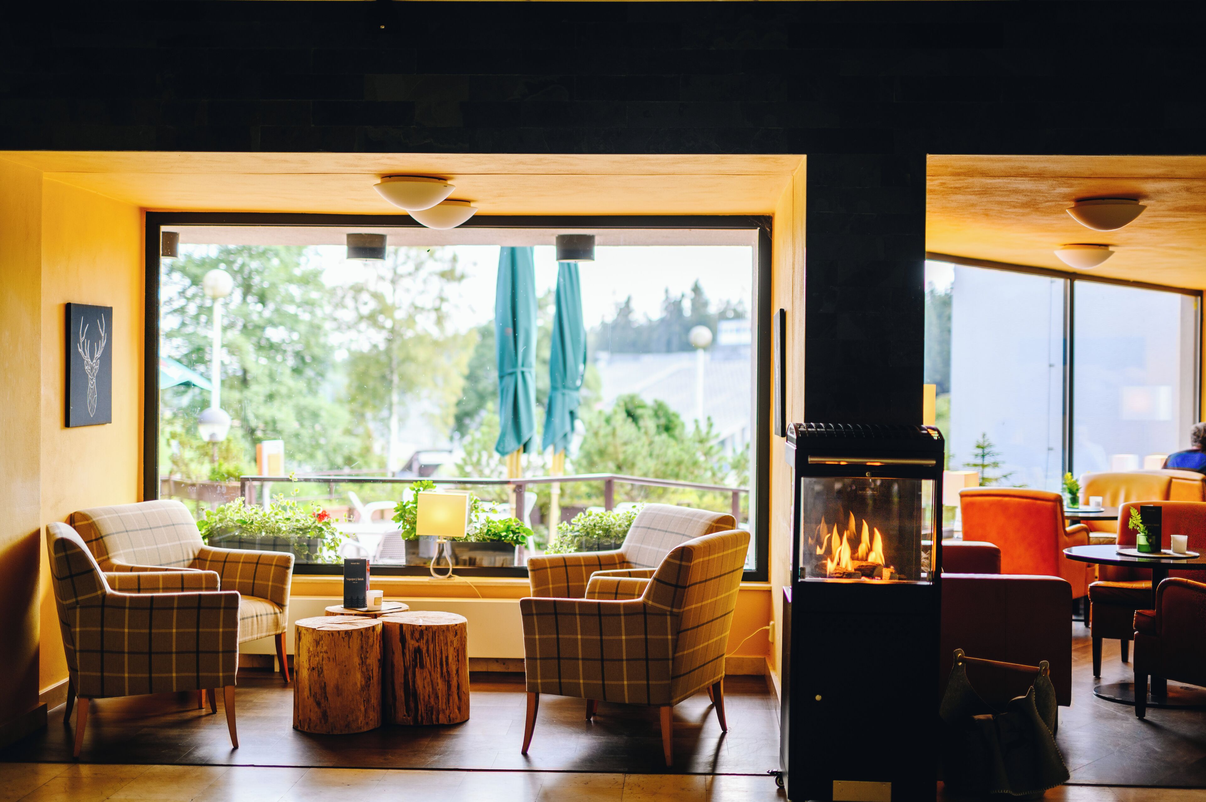 A room with a fireplace, two plaid chairs, a round wooden table, and a view of greenery outside.