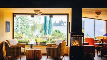 A room with a fireplace, two plaid chairs, a round wooden table, and a view of greenery outside.