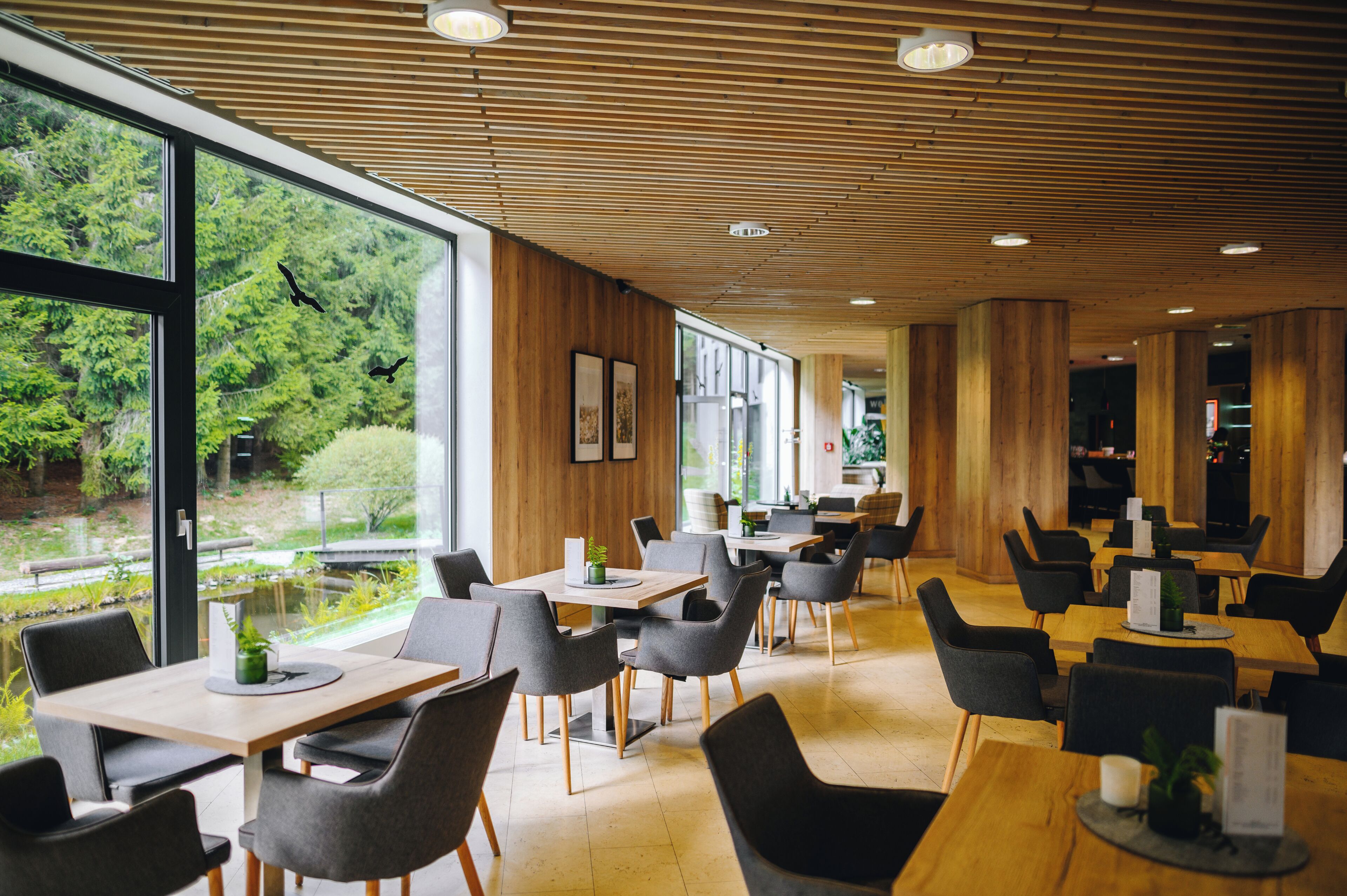 Un ristorante moderno con pannelli in legno a soffitto, ampie finestre con vista sul verde e diverse opzioni di posti a sedere.