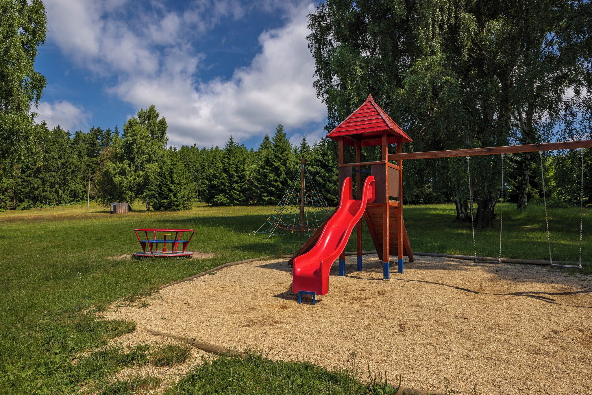Une aire de jeux avec un toboggan rouge, une balançoire et une petite structure en bois.