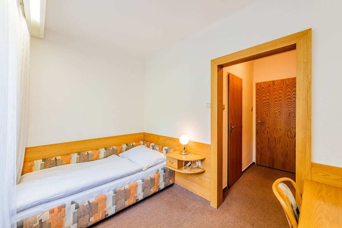 A small, single-bed room with a wooden headboard, a bedside table with a lamp, and a door leading to another room.