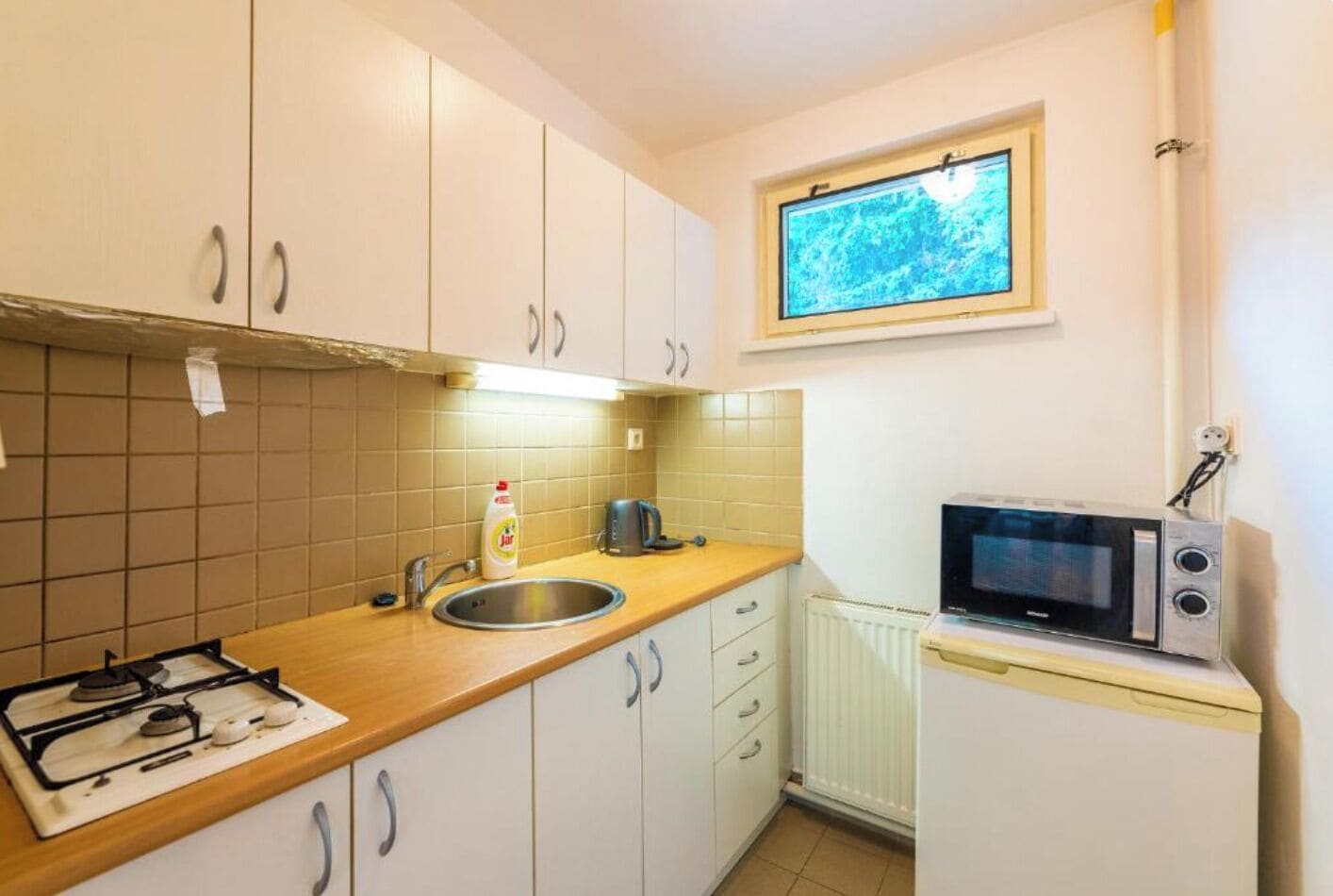Een compacte keuken met een gasfornuis, spoelbak, magnetron en een raam boven het aanrecht.