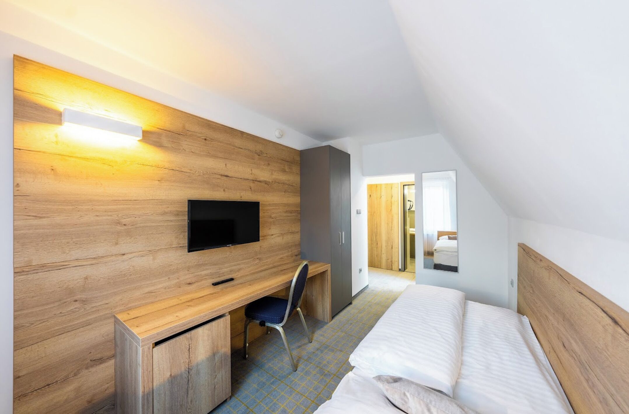 A modern hotel room with a wooden wall, a flat-screen TV, a desk with a chair, and a bed.