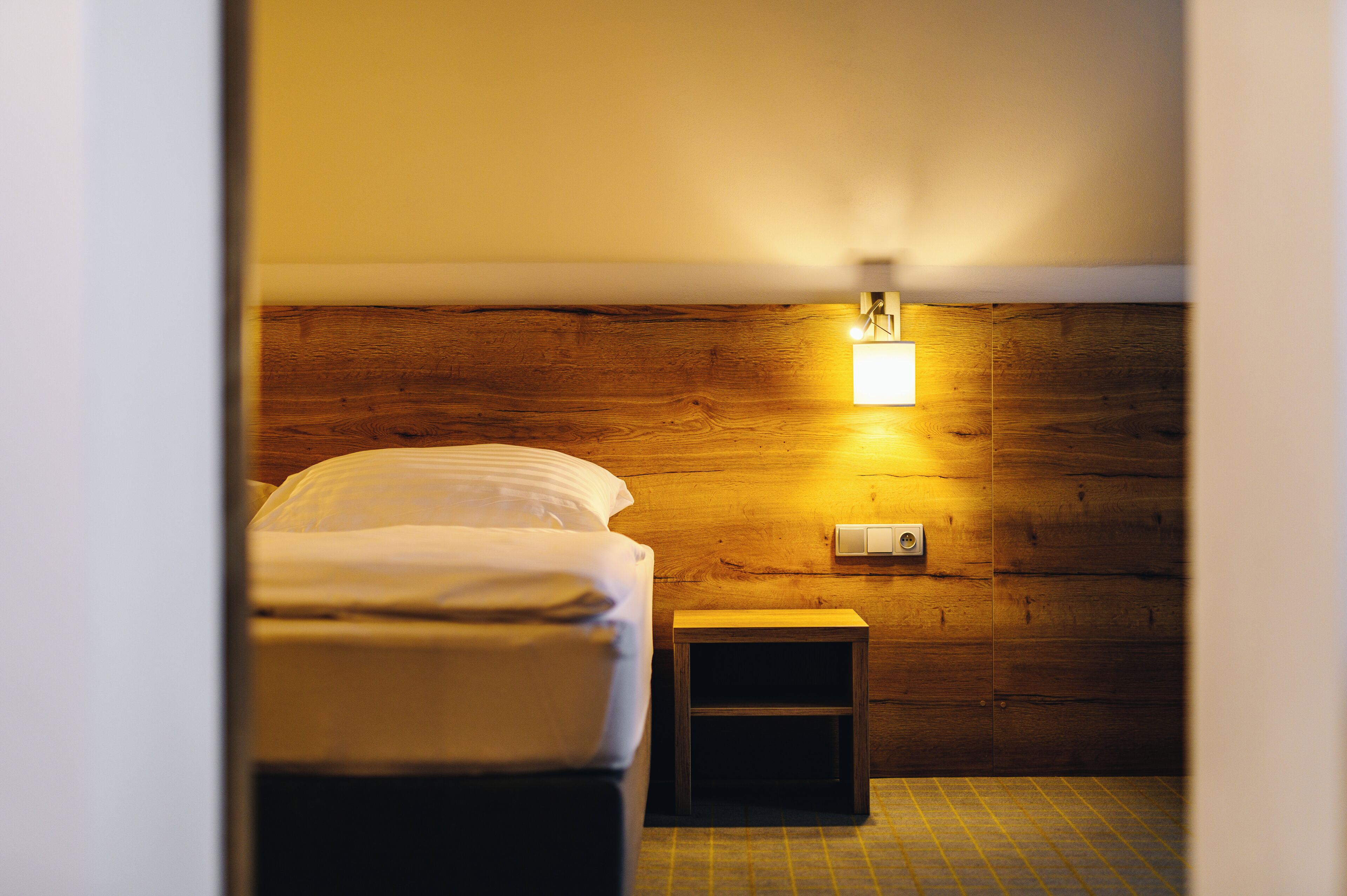 Une petite pièce peu éclairée, avec un mur en bois, un lit simple, une table de chevet et un luminaire fixé au mur.