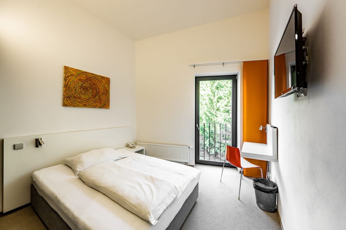 A modern hotel room with a bed, a TV mounted on the wall, a window with a view of greenery, and a door leading to an outdoor area.