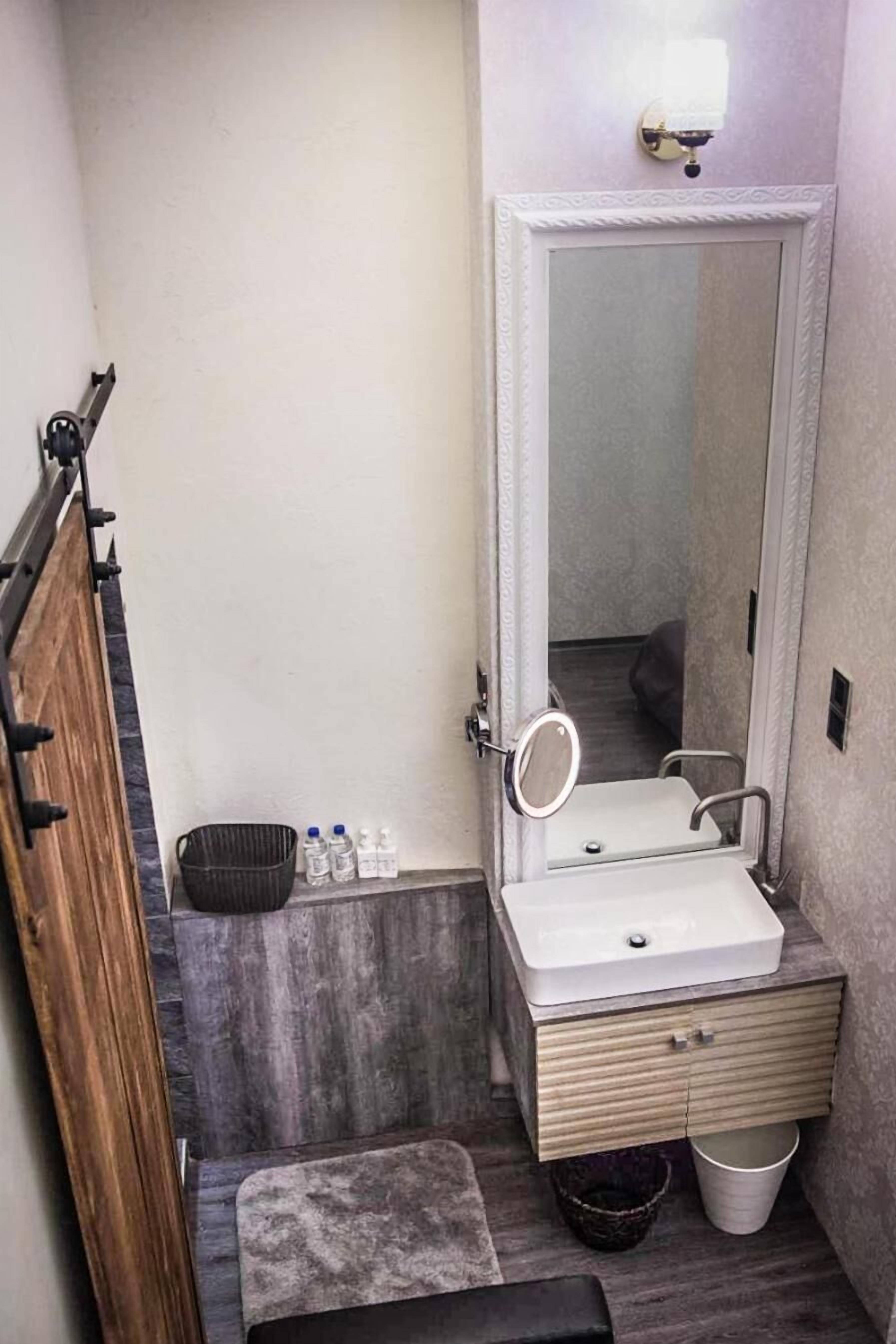 A compact bathroom with a white sink, a mirror, a wooden bench, and a small trash bin.