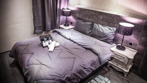 A neatly made bed with a grey duvet, two bedside tables with lamps, a small wooden chest of drawers, and a basket of toiletries. - Summer night (Tainan)