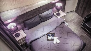 A neatly made bed with a purple comforter and pillows, two bedside lamps, and a small tray with toiletries on the nightstand. - Summer night (Tainan)