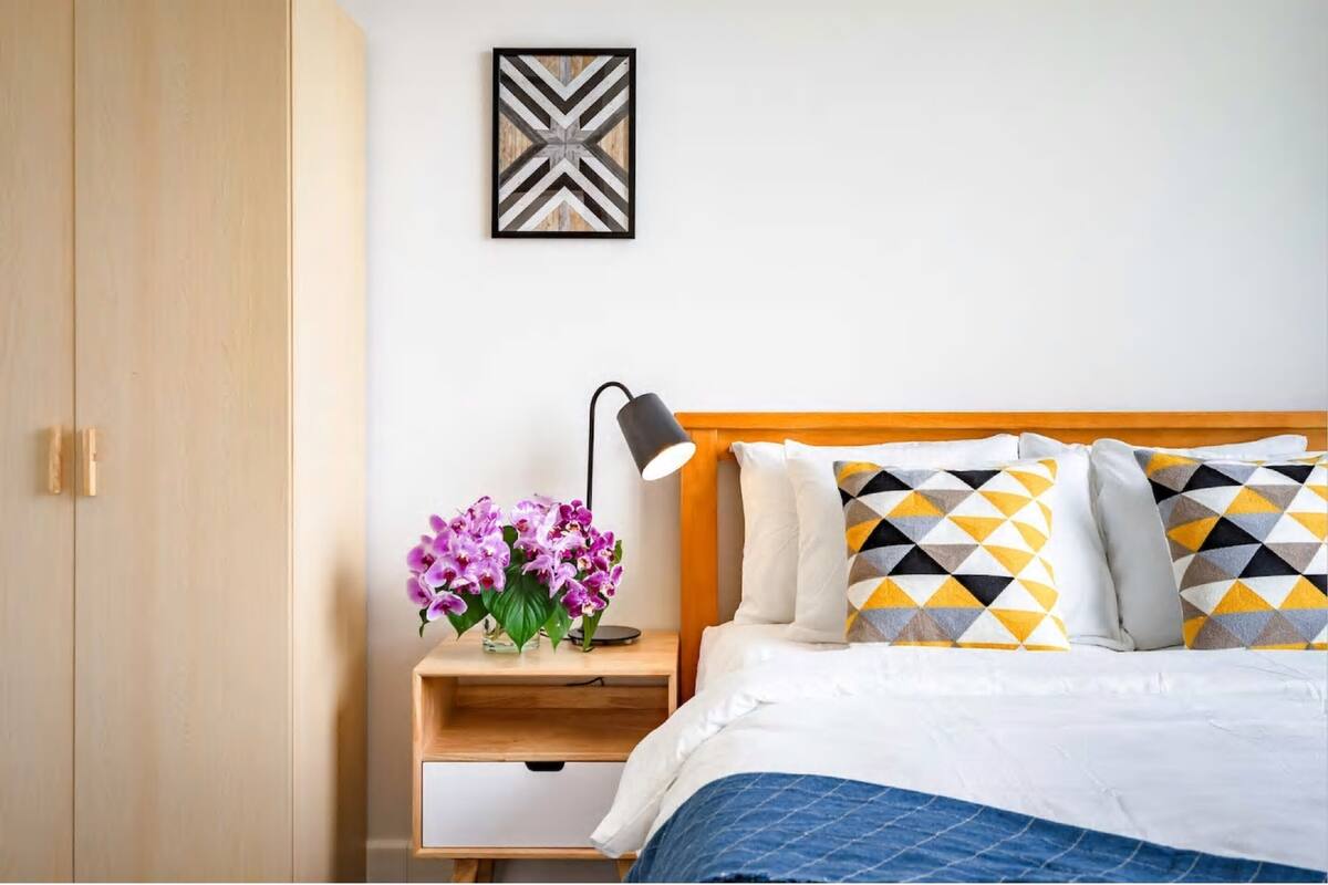 Une chambre avec une armoire en bois, une table de chevet avec une lampe, un lit avec des coussins à motifs et un tableau encadré au mur.