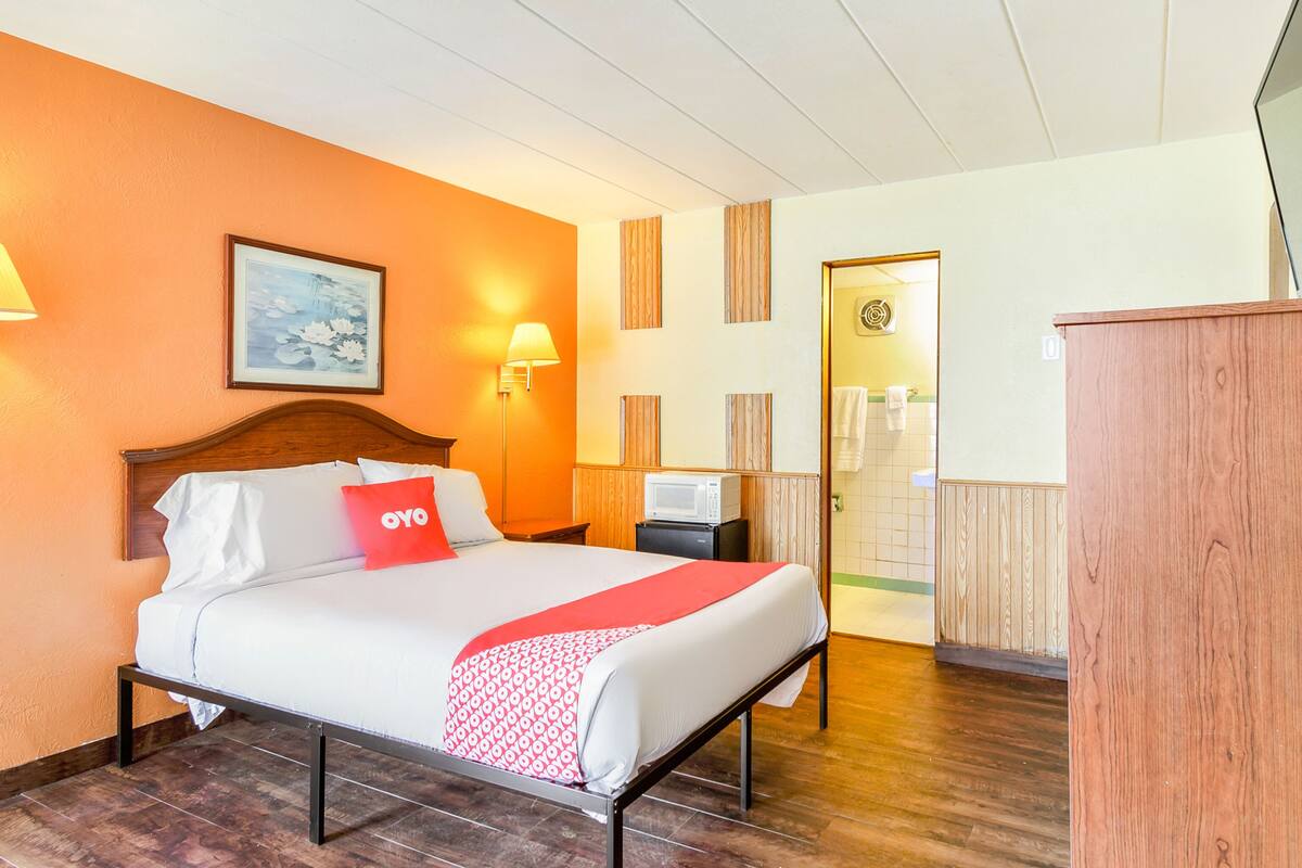 Una habitación de hotel con cama, escritorio, lámpara, un cuadro en la pared y una puerta que da al baño.