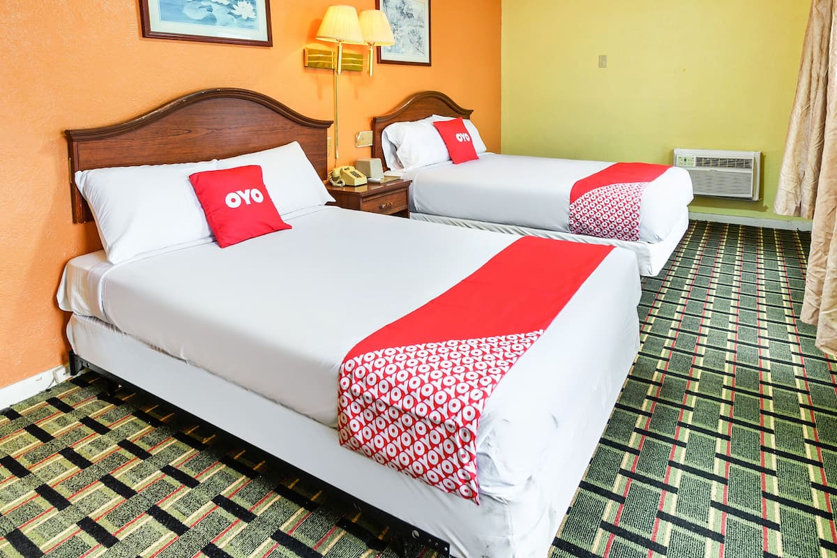 Dos camas con sábanas blancas y detalles rojos, una mesita de noche con lámpara y una alfombra estampada.