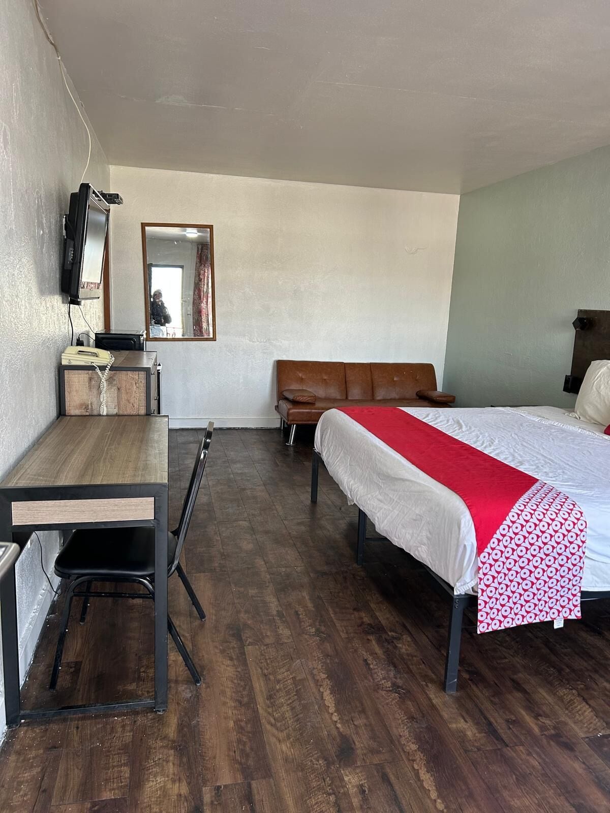 Ein Zimmer mit einem Holztisch, einem braunen Ledersofa, einem Bett mit einer rot-weißen Bettdecke, einem Fernseher und einem Spiegel an der Wand.