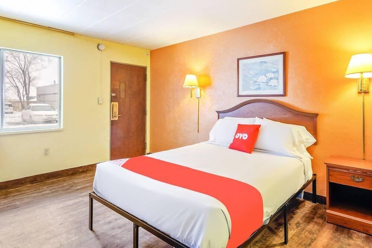 Habitación de hotel con cama, mesita de noche, lámpara de pared, puerta y ventana con vistas a un coche y árboles.