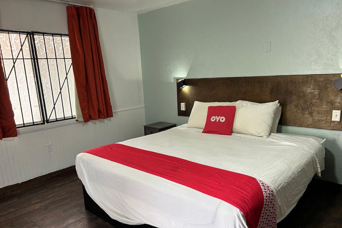 Una habitación de hotel con cama, mesita de noche, ventana con cortinas y cabecero.