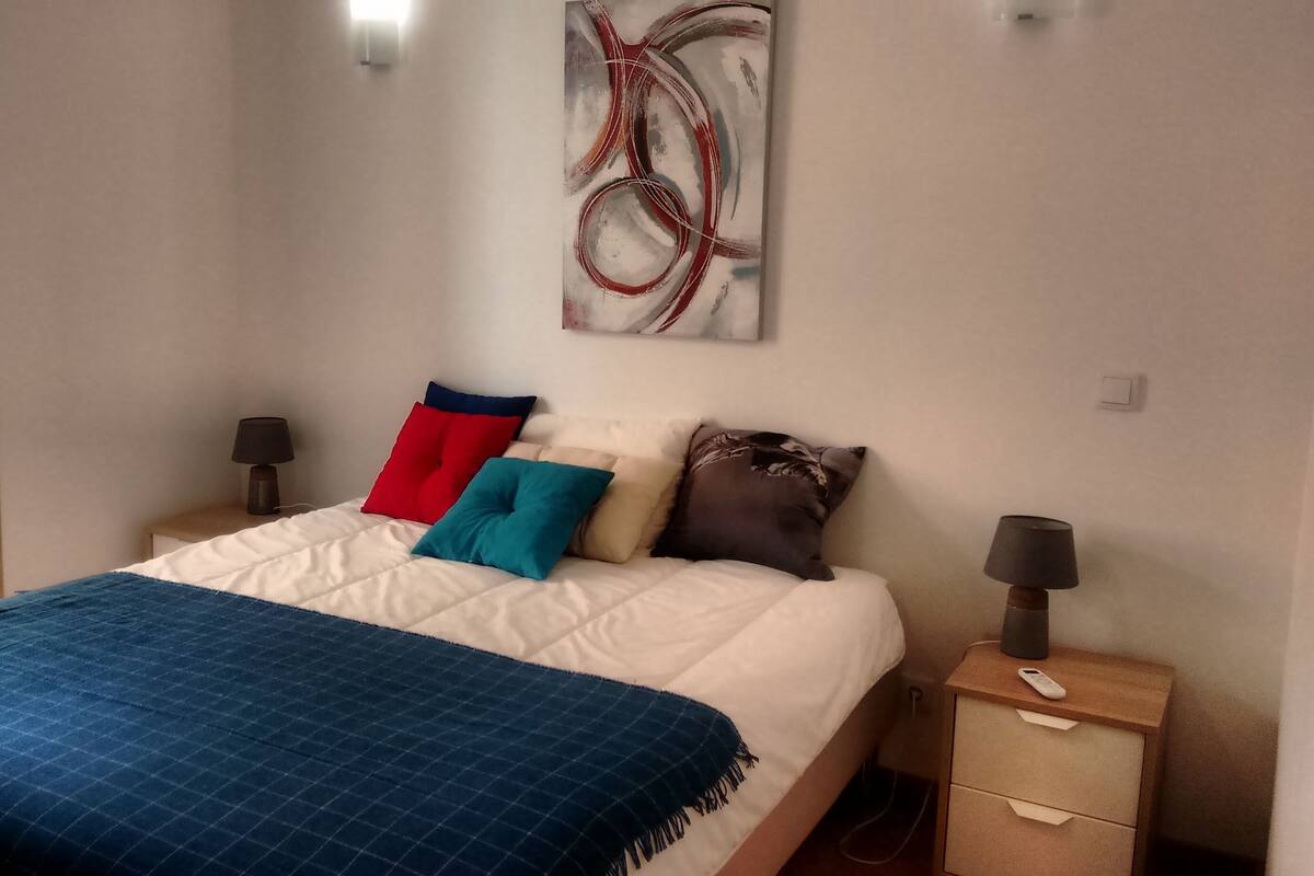 Un dormitorio con cama, almohadas, una mesita de noche, una lámpara y un cuadro en la pared.