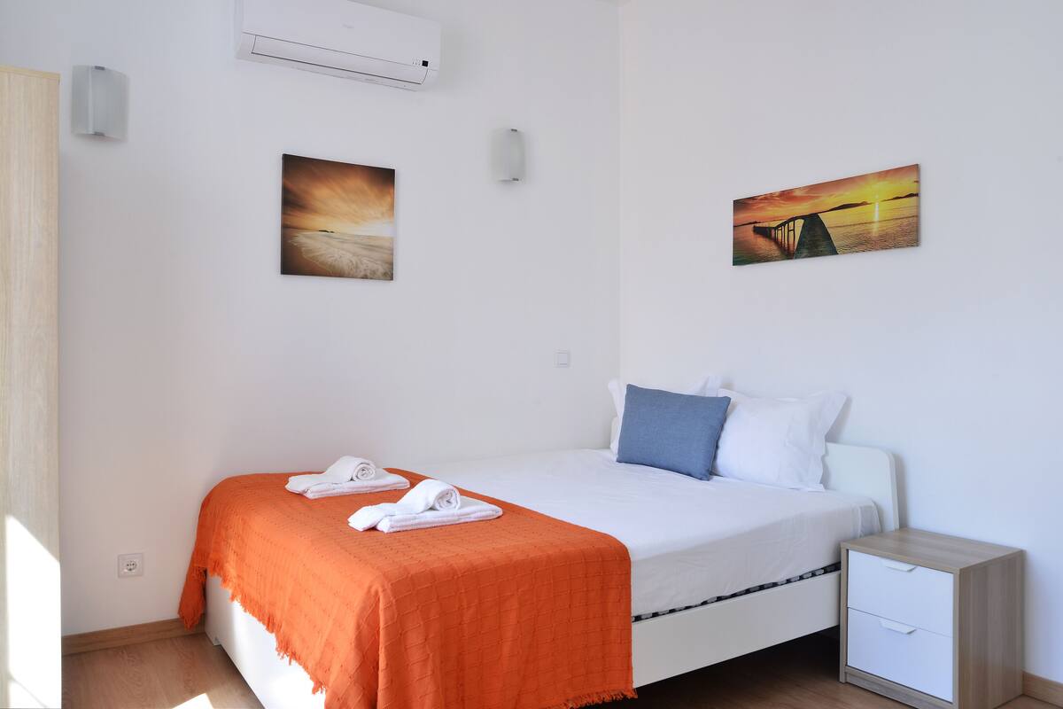 Una cama bien hecha con un edredón naranja, dos almohadas y toallas enrolladas. Hay dos lámparas fijadas a la pared y dos cuadros enmarcados.