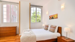 A bedroom with a bed, wooden dresser, nightstand, and a window with a view of buildings and trees. - Oeiras  Apartment (Oeiras)