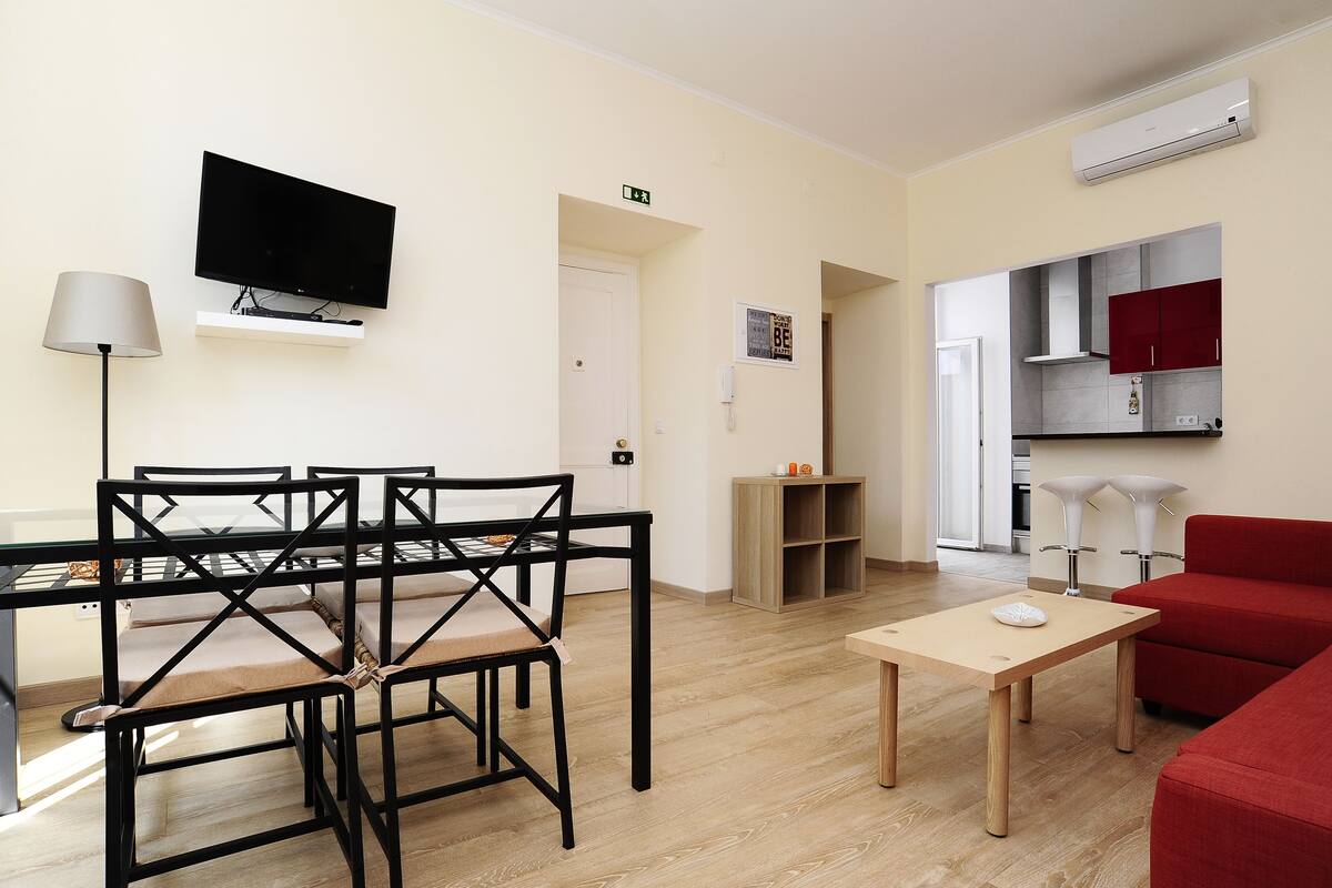 Una sala moderna con comedor, un sofá rojo, un televisor montado en la pared y una cocina visible a través de una puerta abierta.