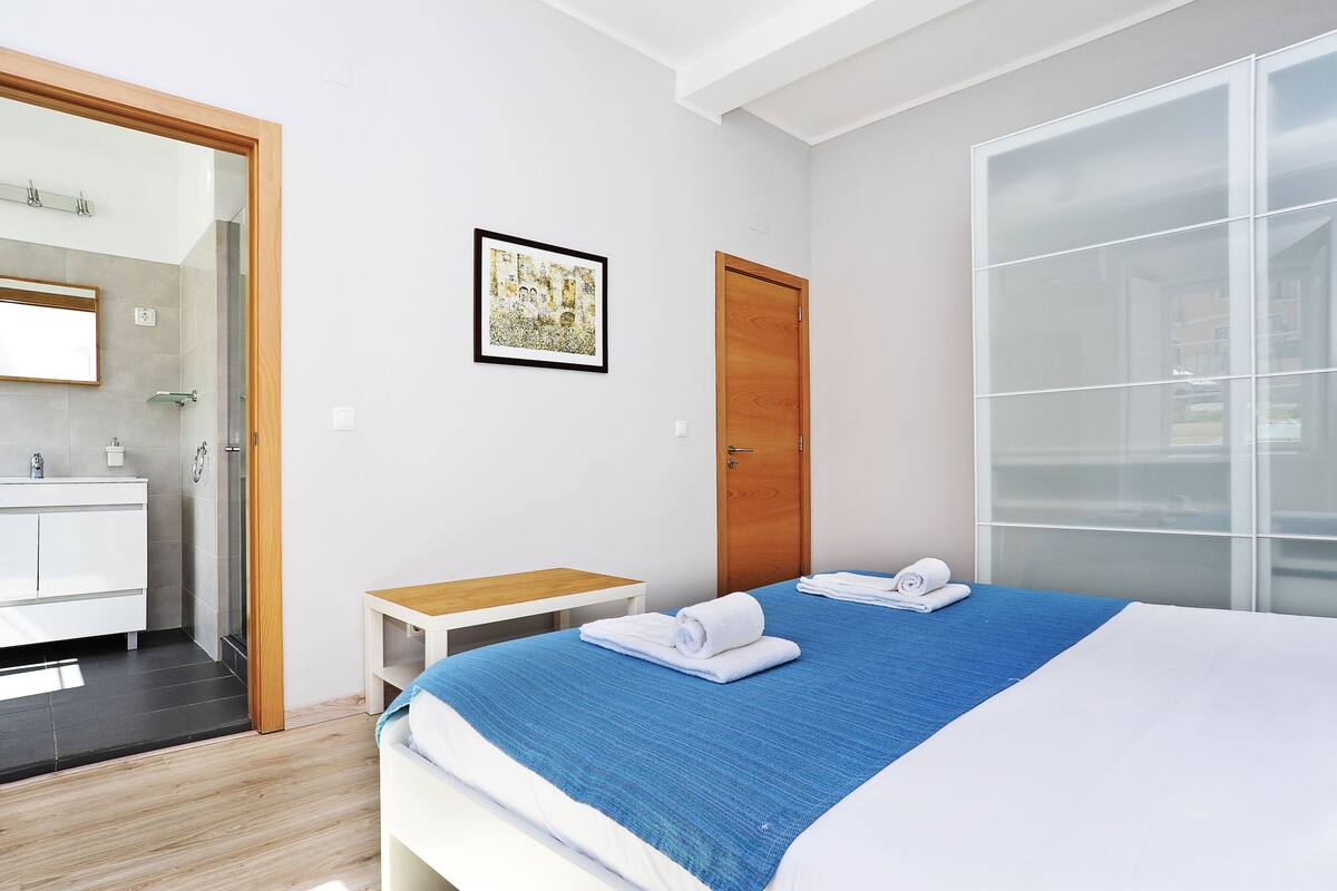 Habitación de hotel moderna con cama, mesita de noche, puerta de madera, baño con espejo y un cuadro en la pared.