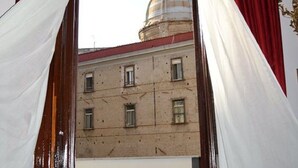 Vista a través de un vano con cortinas rojas, que muestra un edificio con cúpula y un balcón con balaustradas.