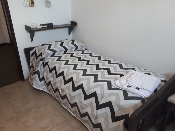 A small, single-bed room with a chevron-patterned bedspread, a bedside table with a lamp, and a wall-mounted power outlet.