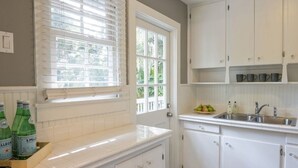 Eine Küche mit weißen Schränken, einem Fenster mit Jalousien und einer Arbeitsplatte mit Flaschen.