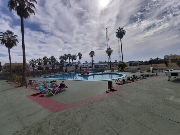 A pool area with lounge chairs and a pool table, surrounded by palm trees and buildings.